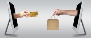 Two computer screens with human hands extended one holding a shopping bag and the other a bank card showing ecommerce transaction taking place 